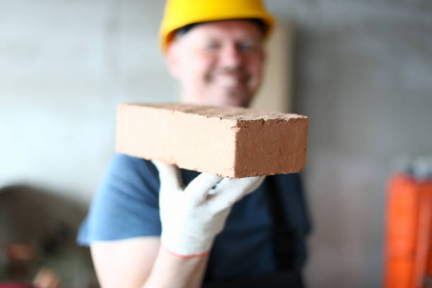Prudent builder in gloves Focus on serious brick in male hand. Worker in hardhat presenting red block to camera with big smile. Joyful constructor in yellow helmet looking with happiness on face. Building concept stacker stock pictures, royalty-free photos & images
