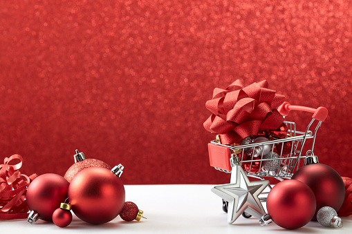 Conceptual photo of Supermarket trolley filled with Christmas sales decorations on a background of holiday red lights. Preparing for the New Year and Christmas. Buying gifts.