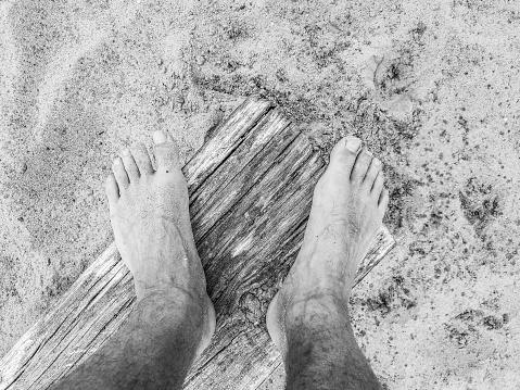 Closeup black and white view looking down at a mans bare feet and toes standing on a piece of driftwood plank or beam on a sandy beach.