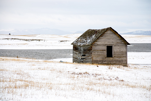In abandoned, wooden homestead structure in a snow-covered field in front of a pond not yet frozen over