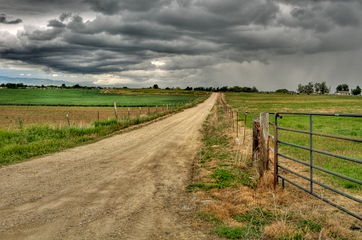 A long dirt road leading off into the horizon past a open fence and into a cloudy and gray stormy sky near a green and grass covered pasture