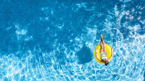 Active young girl in swimming pool aerial top view from above, child relaxes and swims on inflatable ring donut and has fun in water on family vacation, tropical holiday resort