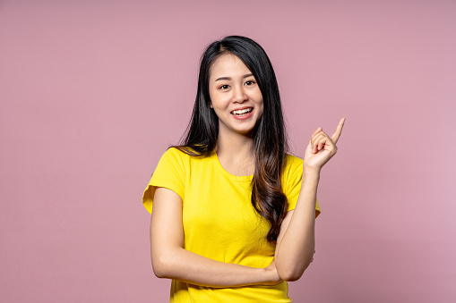 Cute happy asian woman in yellow casual shirt looking at camera, smiling and pointing finger to side on pink studio background for texting or advertise.