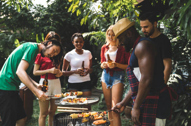 Group of people standing around barbecue grill, chatting, drinking and eating at summer outdoor party and holidays concept. stock photo