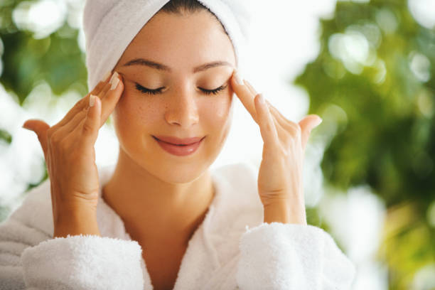 I take care of my skin regularly. Young woman applying and rubbing anti age cream with her eyes closed. She's in the bathrobe and her hair is wrapped in the towel. face cream stock pictures, royalty-free photos & images