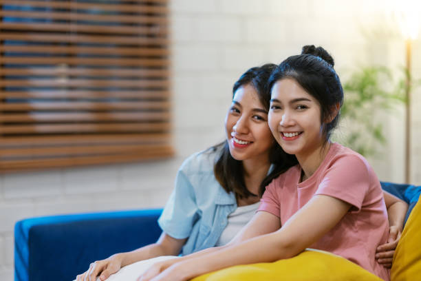 pares asiáticos do com de lgbtq que sorriem, olhando a câmera e sentando-se no sofá na sala de visitas na casa. mulher asiática e sentimento do amigo felizes no tempo da noite. conceito lgbt e amizade. - bff - fotografias e filmes do acervo