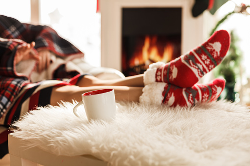 Cut out of female feet on Christmas socks resting on a white carpet near a fireplace.