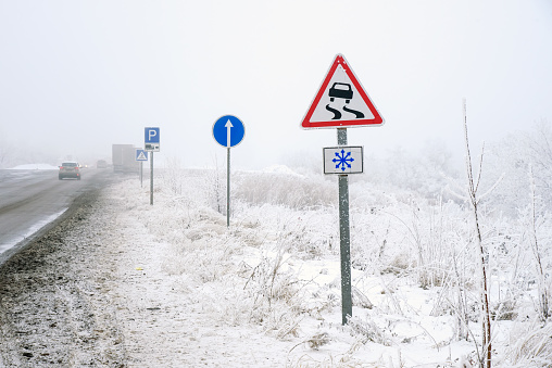 Danger trip on winter road with fog and snow blizzard. Road sign of slippery road, direction of travel, parking.