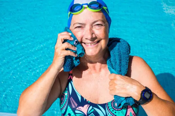 An elderly woman who enjoys the swimming pool. One people with large smile. Blue swimming cap and goggles. Healthy lifestyle by doing physical activity. Sunny day and transparent water