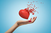 Close-up of woman's hand facing up and levitating red valentine heart that has started to disintegrate into pieces on light-blue background.