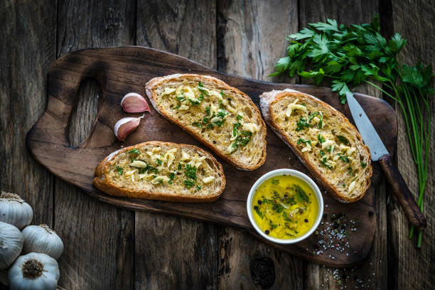 Appetizer: garlic bread on rustic wooden table Appetizer: garlic bread on rustic wooden table. Top view of three toasted bread slices with garlic and parsley are on a wooden cutting board. Parsley bunch, garlic bulbs and olive oil complete the composition. Predominant colors are yellow and brown. XXXL 42Mp studio photo taken with SONY A7rII and Zeiss Batis 40mm F2.0 CF garlic bulb photos stock pictures, royalty-free photos & images