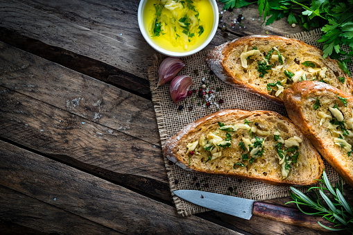 Appetizer: garlic bread on rustic wooden table. Three toasted bread slices with garlic and parsley are on a burlap piece. Parsley bunch, garlic bulbs and olive oil complete the composition. The composition is at the right of an horizontal frame leaving useful copy space for text and/or logo at the left. Predominant colors are yellow and brown. XXXL 42Mp studio photo taken with SONY A7rII and Zeiss Batis 40mm F2.0 CF