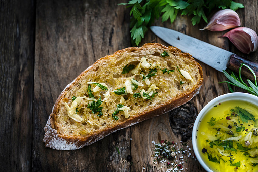 Appetizer: garlic bread on rustic wooden table. Close up view of a toasted bread slice with garlic and parsley shot on rustic wooden table. Parsley bunch, garlic cloves and olive oil complete the composition. Predominant colors are yellow and brown. XXXL 42Mp studio photo taken with SONY A7rII and Zeiss Batis 40mm F2.0 CF