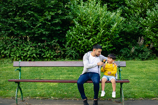 A front-view shot of a cute young boy sitting on a park bench with his dad, they are enjoying ice lollies together.
