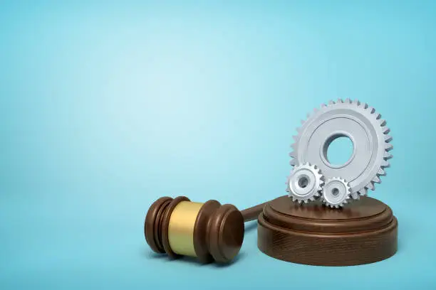 Photo of 3d rendering of one big and two small light-grey gear wheels on sounding block with gavel lying beside on light-blue background with copy space.