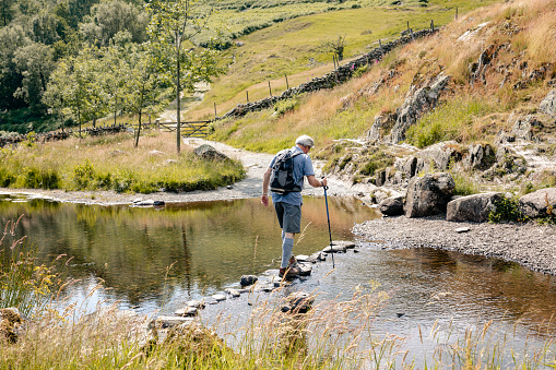 Senior male out hiking crossing river on his own with hiking pole.