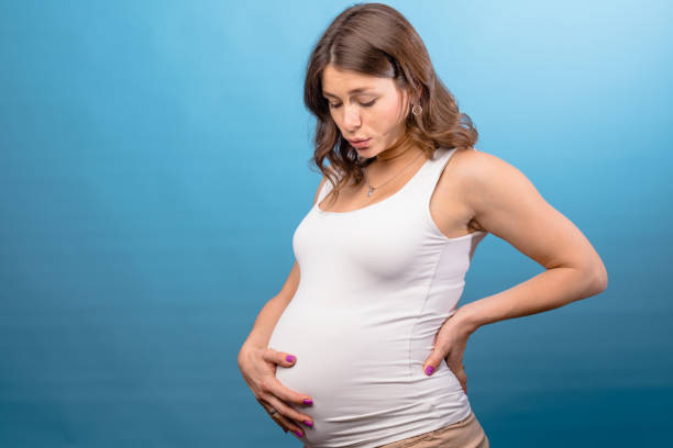 Pregnant woman holding her belly studio shot on pink background Young pregnant woman wearing white shirt holding her belly while keeping one hand on her back suffering from backache. Studio shot on blue background symptom photos stock pictures, royalty-free photos & images