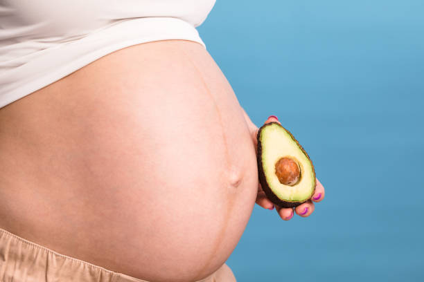 беременная женщина, держащая половину авокадо рядом с животом - плод ягоды стоковые фото и изображения