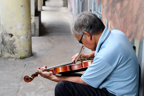 A senior man is sitting against a wall alone and playing violin to past his free time.