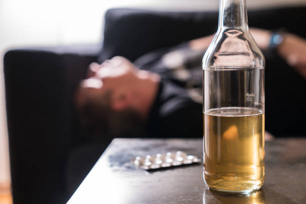 betrunkener mann mit alkoholflasche und pillen auf dem tisch schlafend auf der couch - alcoholism narcotic excess alcohol stock-fotos und bilder