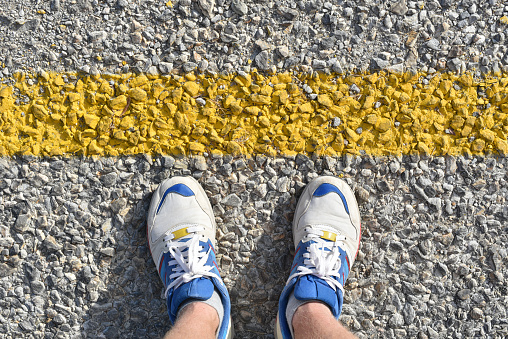 man stands on the asphalt road before yellow border line in white sneakers close-up