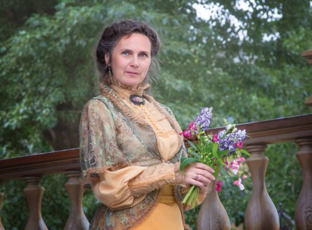 frau in einem historischen kleid auf einer hölzerne veranda mit einem blumenstrauß in den händen. lady teilnehmerin des historischen reenactments - period costume fotos stock-fotos und bilder