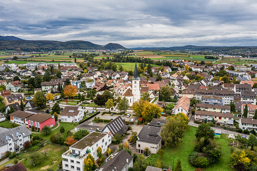 An aerial view of the small town of Therwil in Switzerland. Vivid fall colors of fall foliage against dark dramatic sky. Small colorful houses and churches fill the horizon with mountains in the distance.