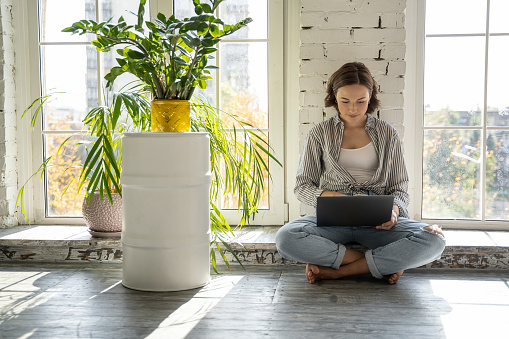 Focused young female freelancer sitting with a laptop on the window sill