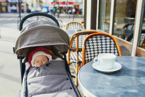 Baby girl sleeping in pram on outdoor terrace of Parisian street cafe with cup of hot coffee on the table. Going out with kids