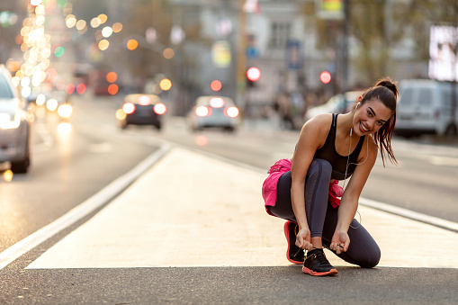 Running woman. Runner is tying laces and listening to music. Female fitness model tying laces outside in the city street, while looking at camera with toothy smile. Sport lifestyle.