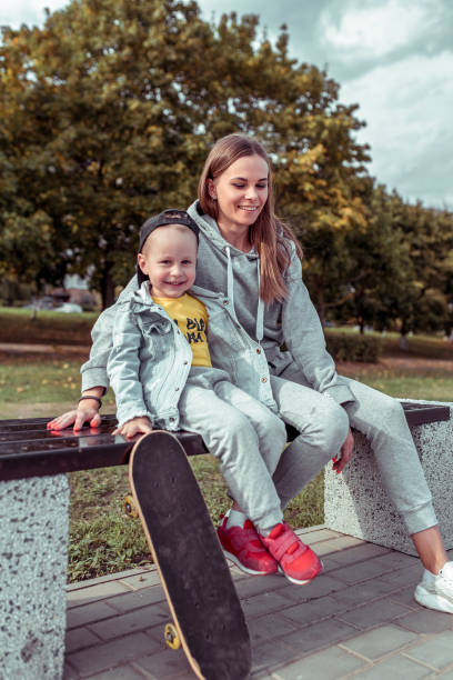 mulher feliz da matriz com filho pequeno 4-5 anos velho, sentando-se no banco no parque da cidade do outono do verão. skate, roupas casuais. descanse em campo de esportes. emoções de alegria e diversão. - child 4 5 years laughing little girls - fotografias e filmes do acervo