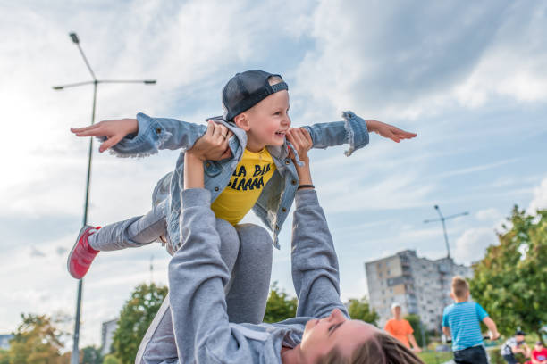 mamá mujer con niño pequeño hijo de 4-5 años de edad, jugar avión, divertirse, reír y disfrutar del descanso, en el sitio del parque de la ciudad de verano. casual use gorra de mezclilla, pantalones de zapatilla. niños de fondo adolescentes. - child 4 5 years laughing little girls fotografías e imágenes de stock