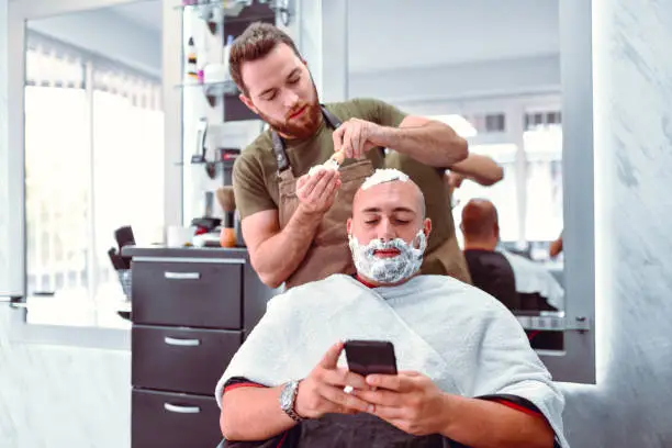 Barber Puts Shaving Cream On Relaxed Customer While He's Using Smartphone