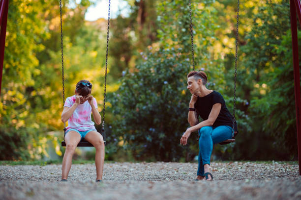 Mother and daughter sitting outdoor on a swing at the playground and having serious conversation