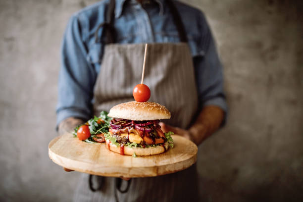 Man with apron standing in the kitchen and holding a wooden plate with prepared delicious hamburgers