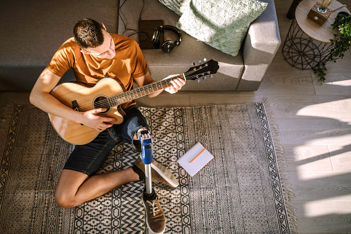 Young man sitting on floor at home and playing acoustic guitar on sunny day