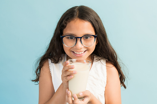 A small joyful girl is at the farm in front of the barn with a glass of milk in her hand and copy space, looking at the camera.