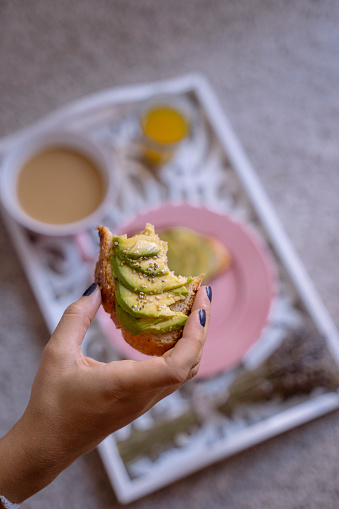 Closeup of women's hands holding an olive oil avocado toast with a fresh bite. Healthy breakfast on a tray. vertical photo.