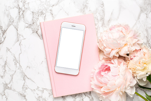 Mobile phone with a white and pink notebook and piony flowers on a marble background top view