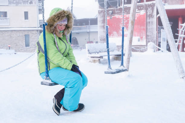 Depression and women's problems. The girl is sitting on a children's swing with her head down. Woman in warm winter clothes, a green jacket and blue pants. Winter and cold outside. Depression and women's problems. The girl is sitting on a children's swing with her head down. Woman in warm winter clothes, a green jacket and blue pants. Winter and cold outside. seasonal affective disorder stock pictures, royalty-free photos & images