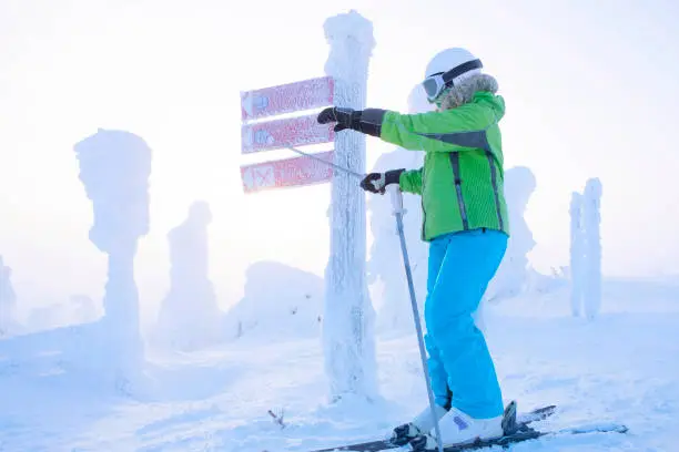 Photo of Girl skier in a green jacket in a helmet and glasses indicates the direction with his hand. Signs of the direction of movement on the ski slopes. Sunrise and beautiful landscape of a snowy forest. Winter holidays in Lapland. Finland.