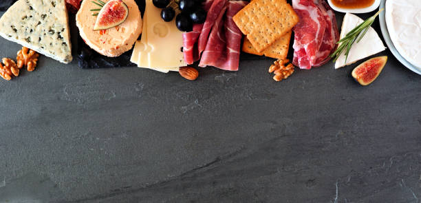 Assorted cheeses and meat appetizers, above view top border banner on a dark stone background with copy space Assorted cheeses and deli meat appetizers. Top border, above view banner on a dark stone background with copy space. cold cuts meat photos stock pictures, royalty-free photos & images