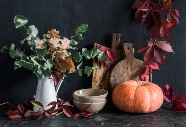 istock Autumn cozy kitchen still life. Crockery, jug with eucalyptus branches, cutting boards, vine wreath, pumpkin on the table, on a dark background. Rustic style. Flat lay 1181986964