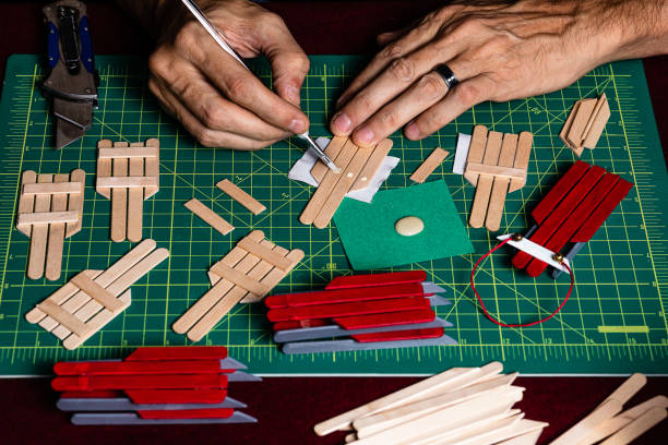 A tightly framed shot of someone working on crafts for the Christmas season. Only the craftsman's hands are visible in the shot while he glues and cuts craft sticks for building Snow Sled Ornaments for a Christmas tree. The background of the image is a bokeh of christmas lights with plenty of space for Copy. Various parts for the craft are spread around the work surface cutting mat. una foto apretada de alguien que trabaja en artesanías para la temporada navideña. sólo las manos del artesano son visibles en la toma mientras pega y corta palos artesanales para la construcción de adornos de trineo de nieve para un árbol de navidad