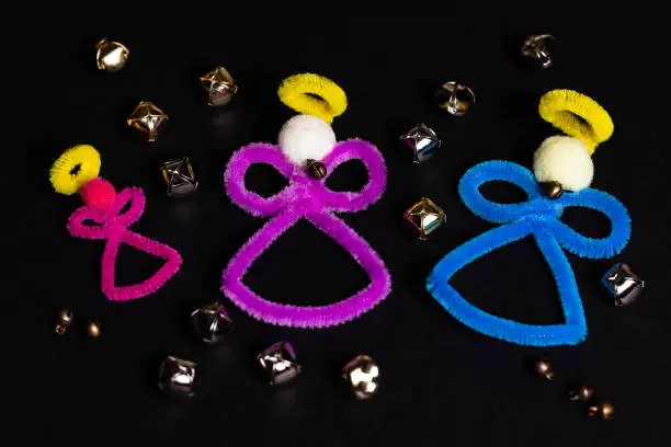 3 pipe-cleaner craft angels on a black background with several gold and chrome jingle bells randomly spread around on the background.  There is plenty of negative space for small copywriting blurbs.  Angels and bells, how an angel gets their wings.