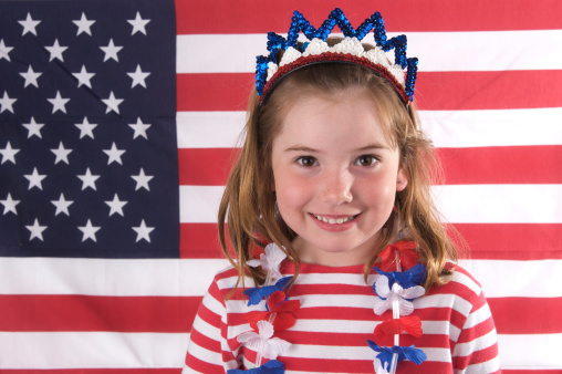A cute little girl is ready for 4th of July with her red white and blue crown and lei.  She's standing in front of the American flag.