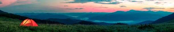 Orange tourist tent illuminated from inside stands in mountains above clouds
