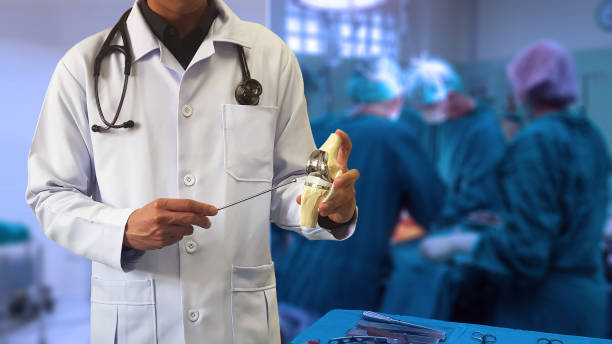 Orthopedic doctor showing model of Total knee replacement with blurred background of surgical team performing surgery in operating room. Medical technology concept. total knee replacement surgery artificial knee photos stock pictures, royalty-free photos & images