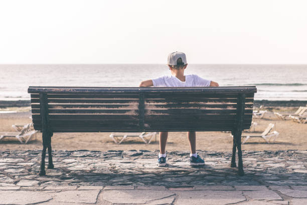 istock Rear View Of Boy Sitting On Bench At Beach 1182037265