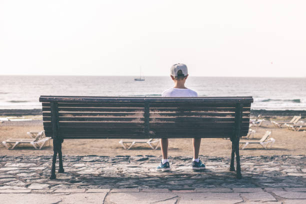 istock Rear View Of Boy Sitting On Bench At Beach 1182038403